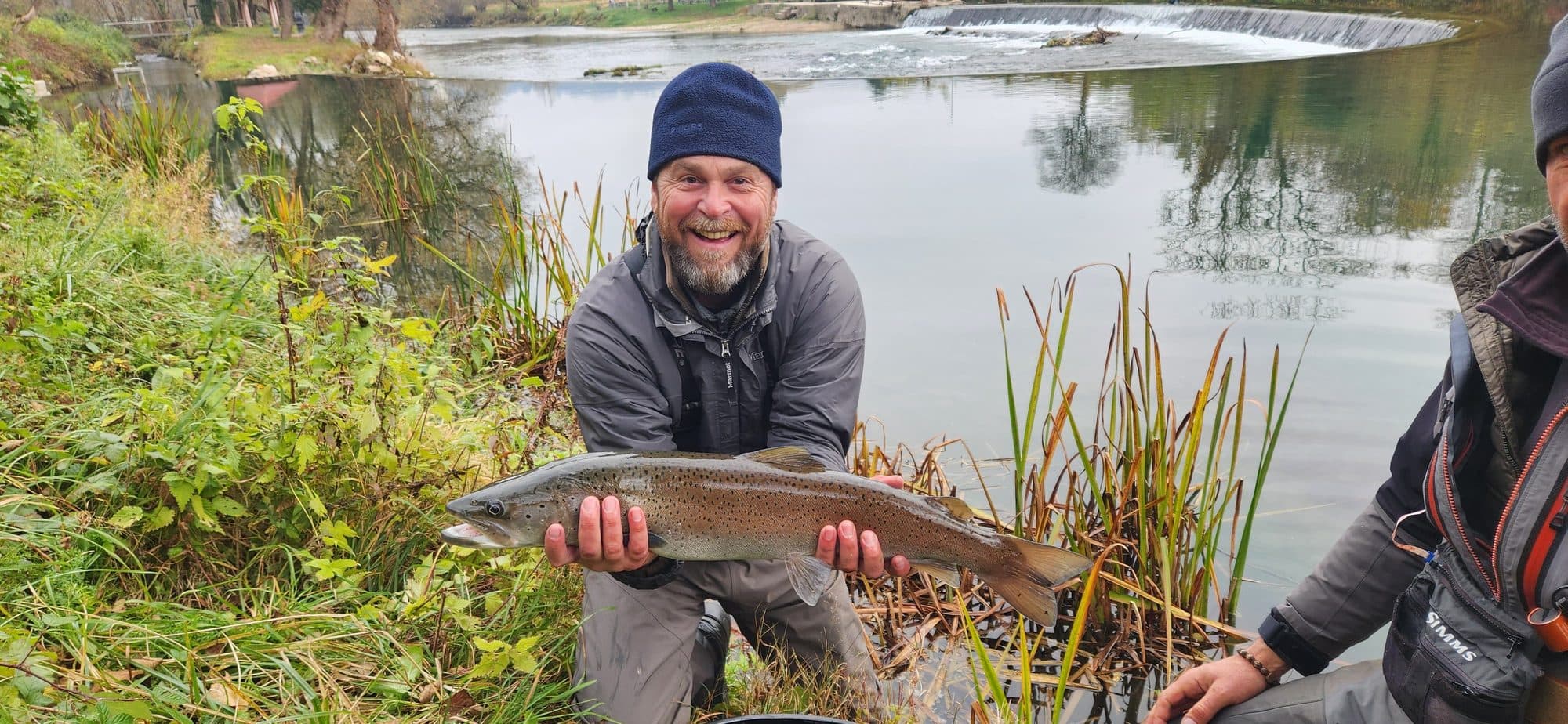 Angler holding a huchen trophy at arm's length beside the river